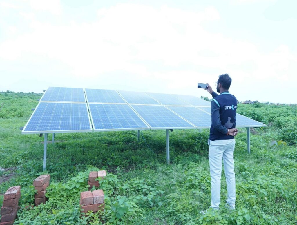 An Anaxee field worker photographs a ground-mounted solar panel array in a lush farm, documenting a solar-agriculture pilot in rural India.