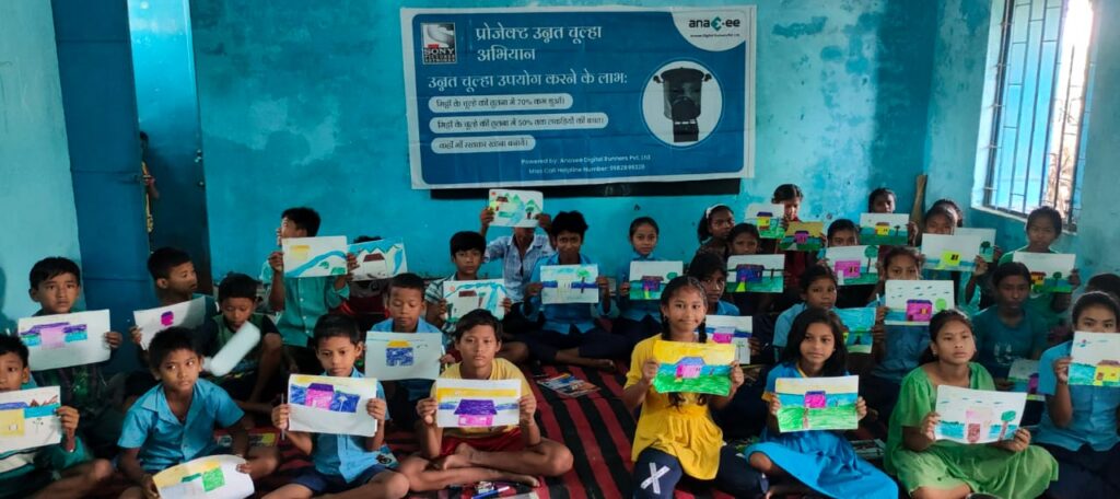 Group of school children in rural India holding colorful drawings during Anaxee’s Project Unnat chulha abhihyan awareness campaign, with a banner about clean cooking solutions displayed behind them.